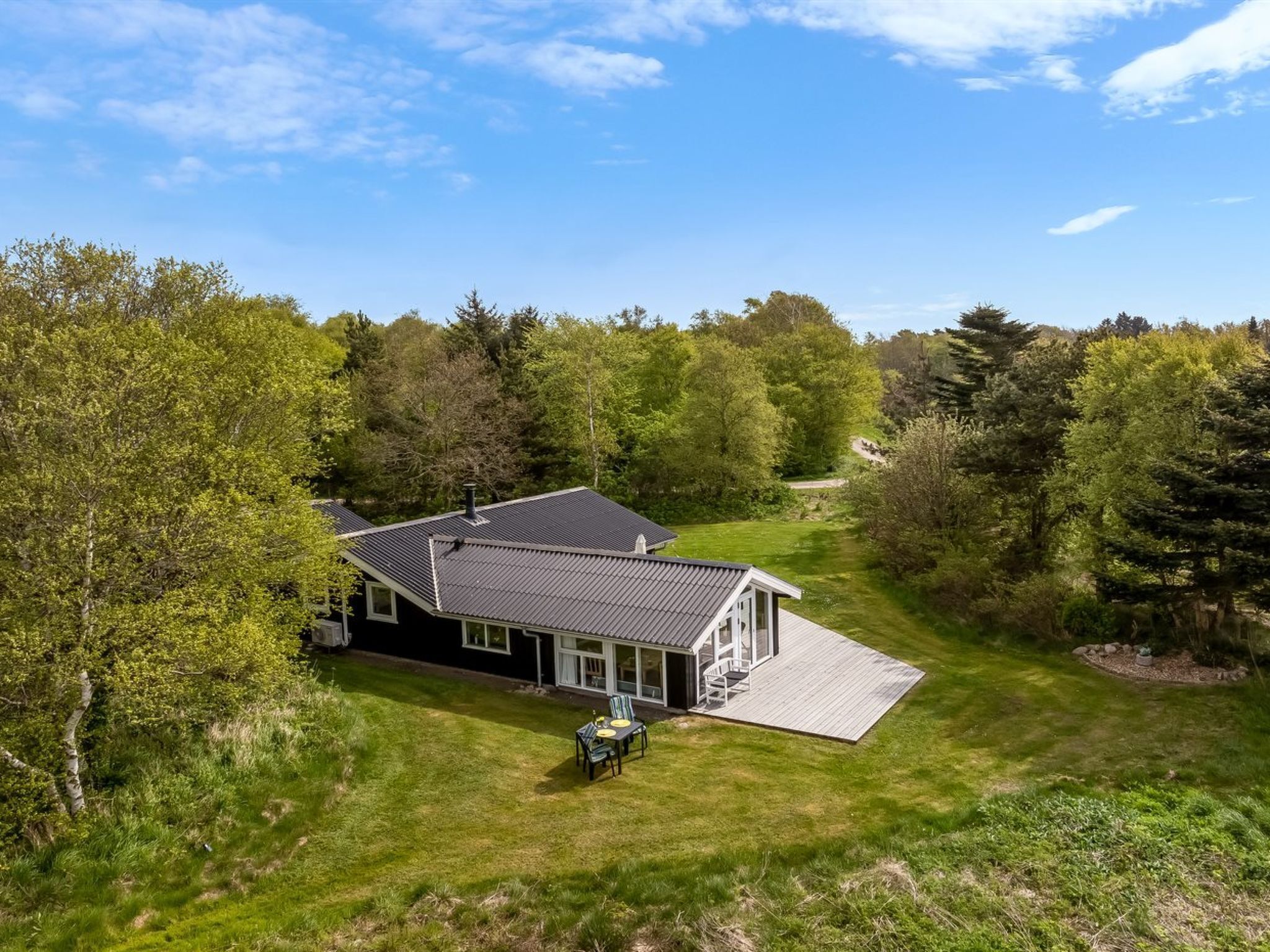 Een huis met een groot terras, omringd door een weelderig groen gazon en dichte bomen.