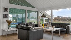 Un salon moderne avec un canapé en cuir noir, une table basse blanche et un mur en verre offrant une vue sur l’extérieur.