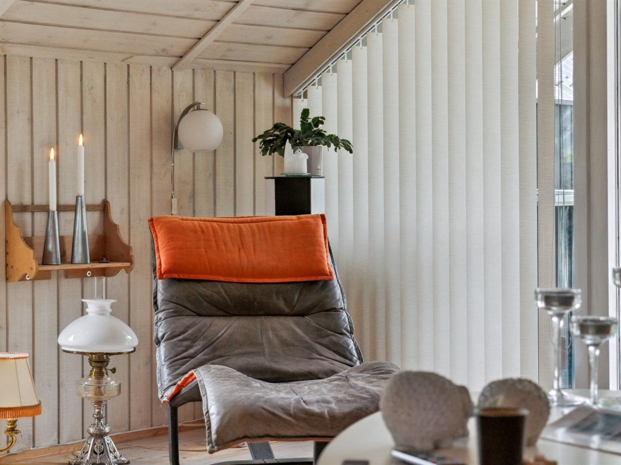 Ein gemütliches Zimmer mit Holzbalkendecke, einem gepolsterten Sessel mit orangefarbenem Kissen, einer weißen Lampe und einer Topfpflanze auf einem Regal.