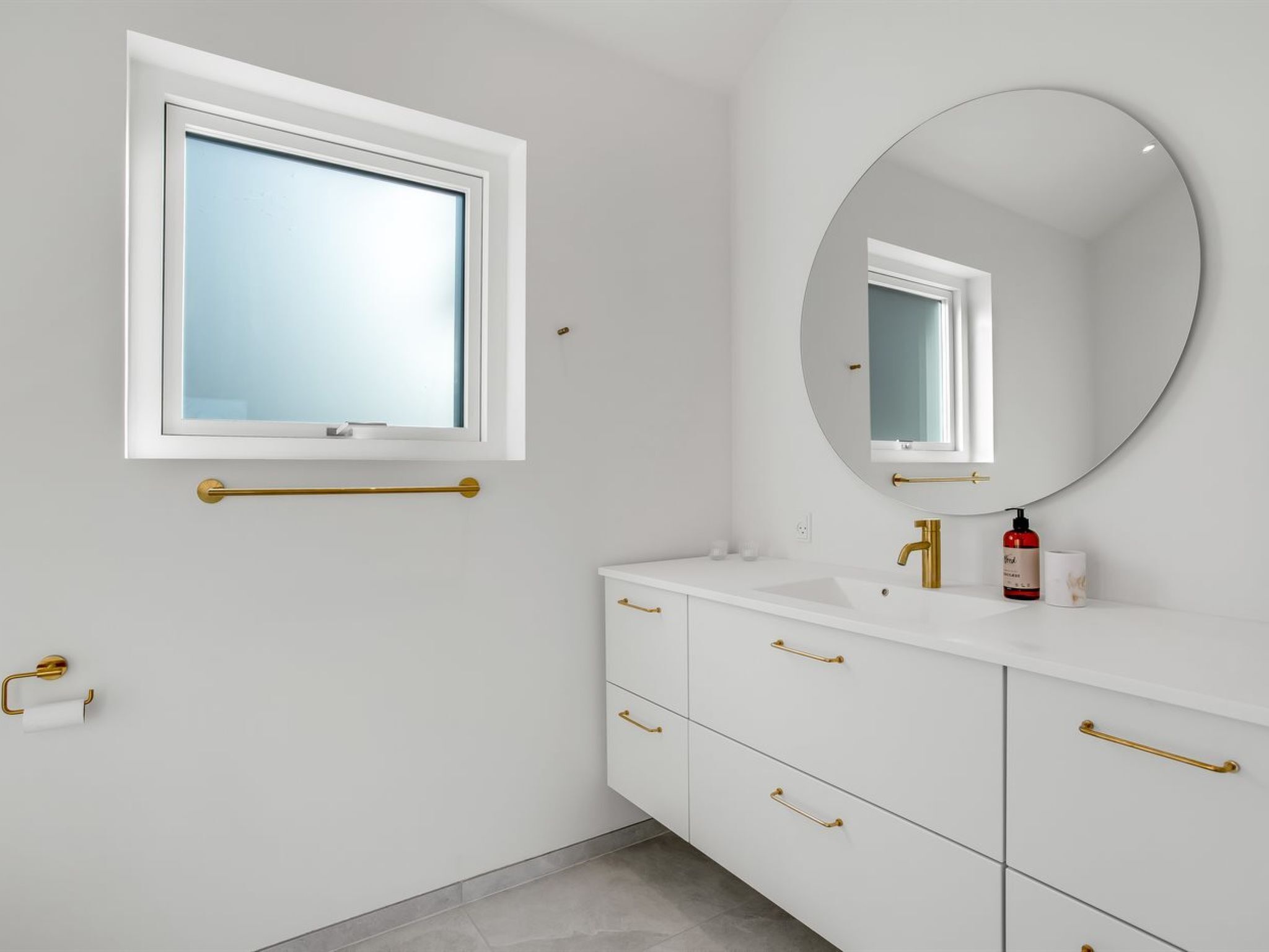 A bathroom with a window, a round mirror, a sink, and a toilet.