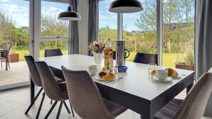 Een ontbijtruimte met een gedekte tafel, inclusief gebak en een kan sap, met grote ramen met uitzicht op de omgeving.