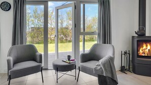 Een moderne woonkamer met twee grijze fauteuils, een klein tafeltje met een boek en een kopje, een open haard en grote ramen met gordijnen.