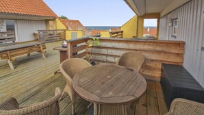 Une terrasse en bois avec une table ronde et des chaises en osier, donnant sur une zone côtière avec des bâtiments et la mer.