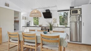 A modern kitchen with a dining area, featuring a large island, wooden chairs, a hanging light fixture, and a stainless steel refrigerator.