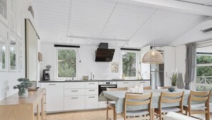 A modern kitchen with white cabinetry, a dining area with wooden chairs, and large windows offering views of greenery.