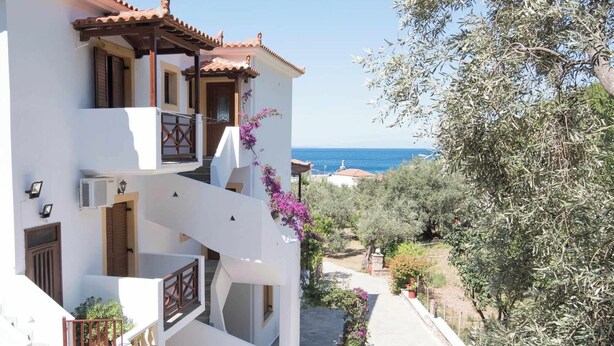 Un bâtiment à deux étages, doté d’une façade blanche, de tuiles en terre cuite et d’un balcon orné de fleurs violettes. On aperçoit une vue dégagée sur la mer à travers le feuillage.
