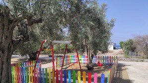 Un terrain de jeux avec des clôtures et des balançoires en bois aux couleurs vives, entouré d’oliviers.