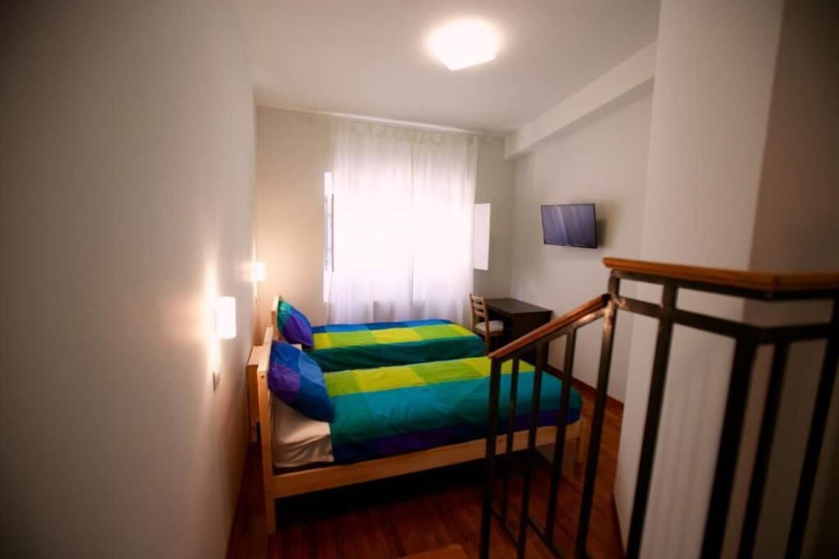 A small, single-bed room with a wooden floor, a desk, a chair, a TV mounted on the wall, and a window with curtains.