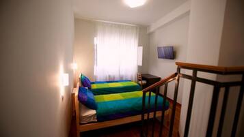 Ein kleines Zimmer mit einem Einzelbett, Holzboden, Schreibtisch, Stuhl, Wand-Fernseher und einem mit Vorhängen versehenen Fenster.