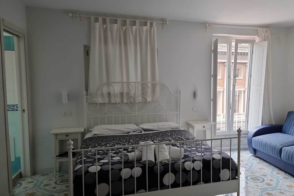 A bedroom with a bed, a nightstand, a window with curtains, and a blue sofa.