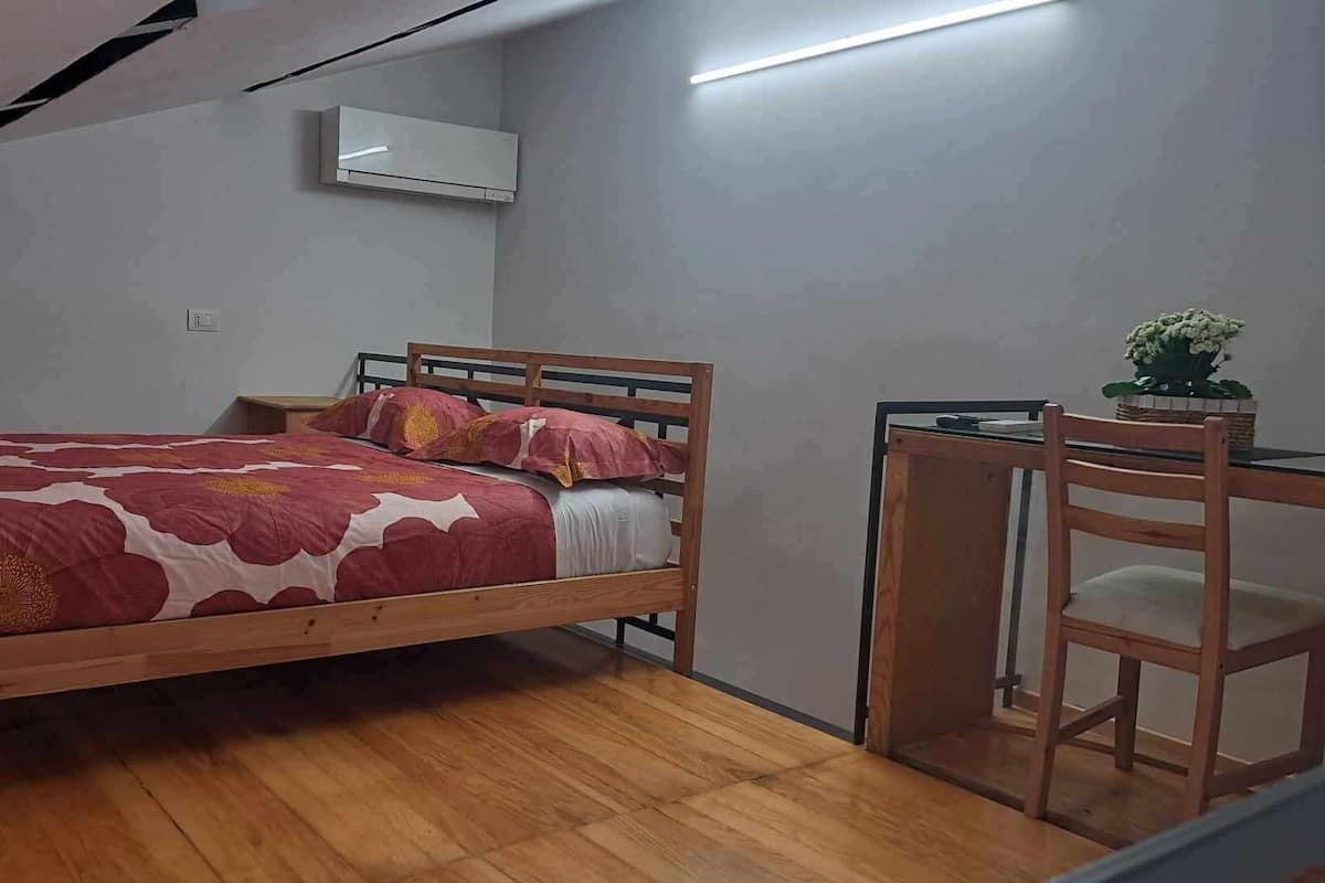 A single bed with a wooden frame and a red floral bedspread, a small table with a chair, and an air conditioning unit on the wall.