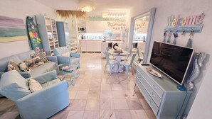 Ein Wohnzimmer mit Fernseher, blauen Möbeln, einem Esstisch mit Stühlen und einer Küche im Hintergrund.