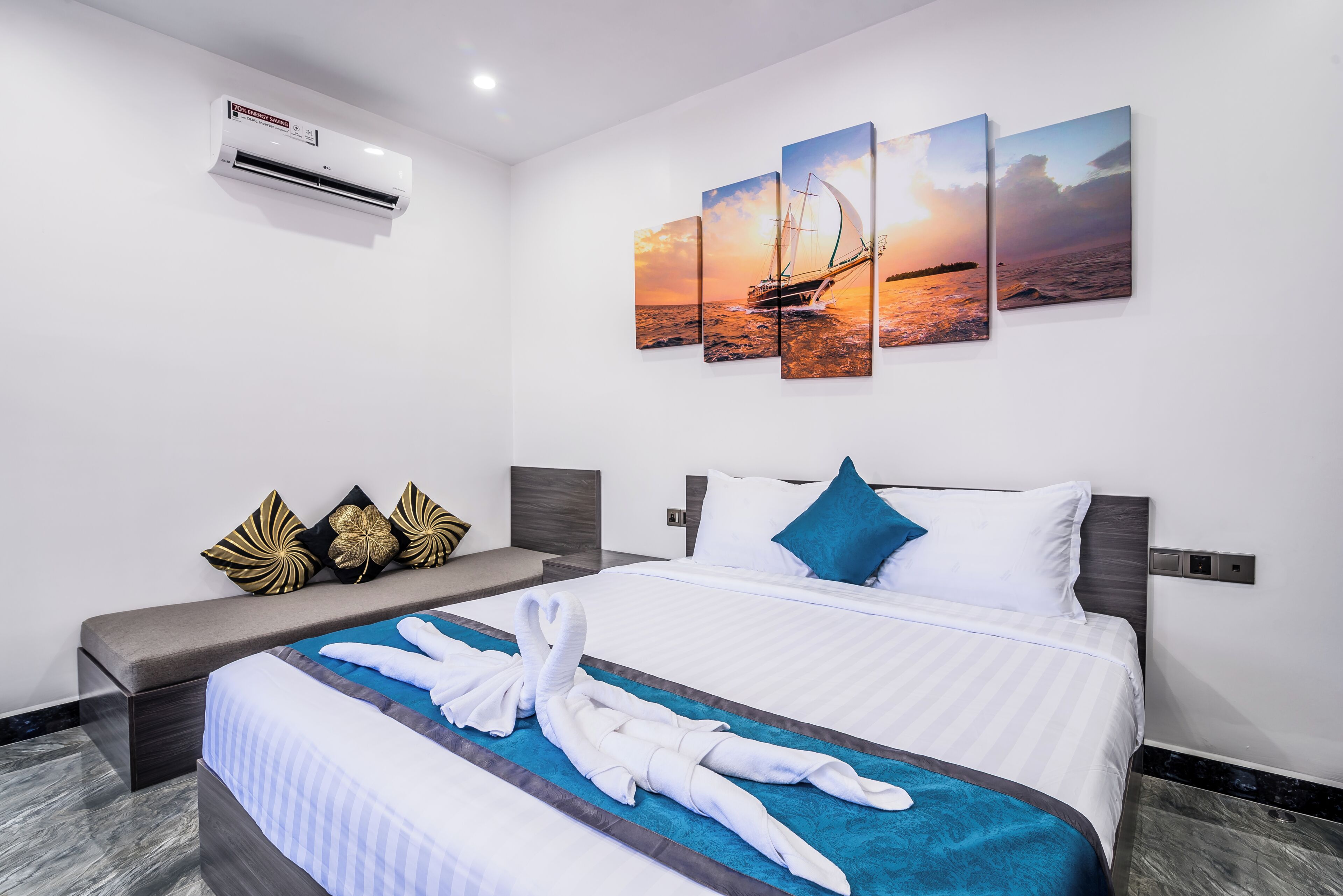 A hotel room with a bed, a wall-mounted air conditioner, and a set of three paintings depicting a sunset over the sea.