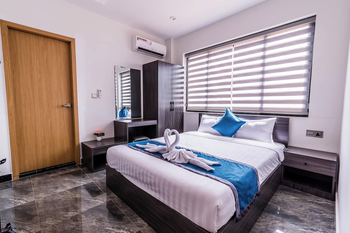 A modern hotel room with a large bed, a wooden door, an air conditioning unit, a bedside table, a nightstand, and a window with blinds.