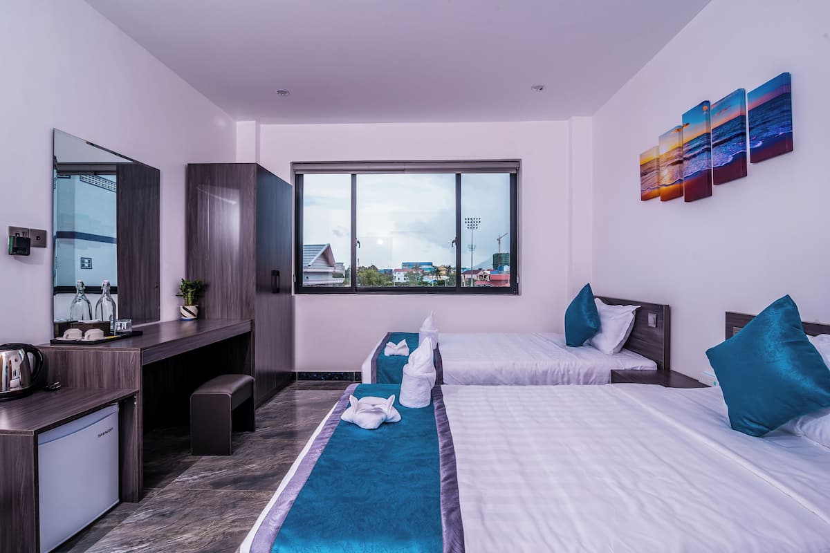 A modern hotel room with two beds, a desk, a small kitchenette, and a large window offering a city view.