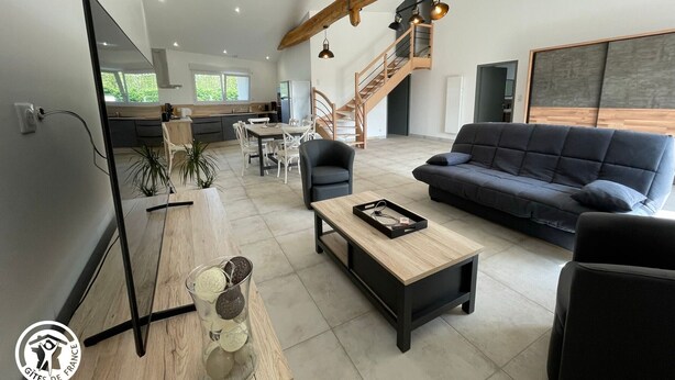 A modern living room with a sofa, coffee table, and TV. There's a staircase with a wooden railing leading to a balcony. - Cozy Cottage for 6 with Terrace – Near La Faute sur Mer & Puy du Fou (Les Magnils-Reigniers)