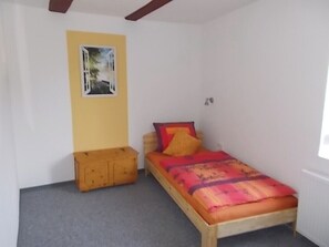 A small, single-bed room with a wooden bed frame, a colorful bedspread, and a small wooden chest of drawers.