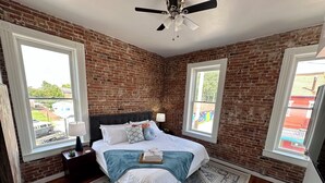 Une chambre avec un mur en briques, un lit recouvert de draps blancs, un plaid bleu et un tapis à motifs.