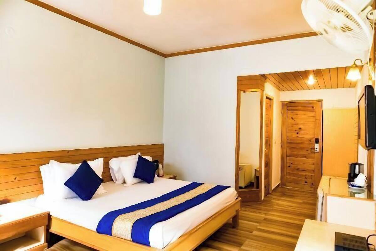 A hotel room with a wooden bed, a desk with a chair, a bedside table, a wall-mounted TV, and a wooden door leading to another room.