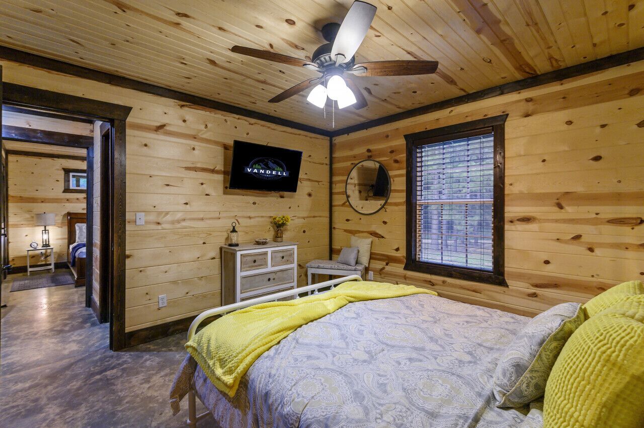 Een slaapkamer met houten lambrisering, een bed, een plafondventilator, een televisie, een raam met zonwering en een klein tafeltje met een lamp.