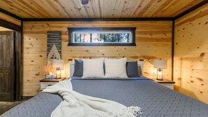 Dormitorio de cabaña de madera con cama, dos mesitas de noche, una ventana y vista a árboles.