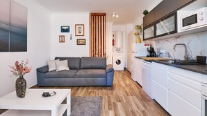 Un espace de vie compact comprenant un canapé gris, une table basse blanche, un coin cuisine avec un four à micro-ondes et un lave-linge.