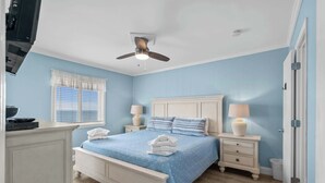 A bedroom with a bed, two bedside tables, a ceiling fan, a window with blinds, and a door. - Duneside Unit 302 - Oceanfront (Murrells Inlet)