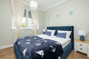 A bedroom with a navy blue bed, white and navy patterned bedding, a white nightstand with a lamp, and nautical-themed wall art.