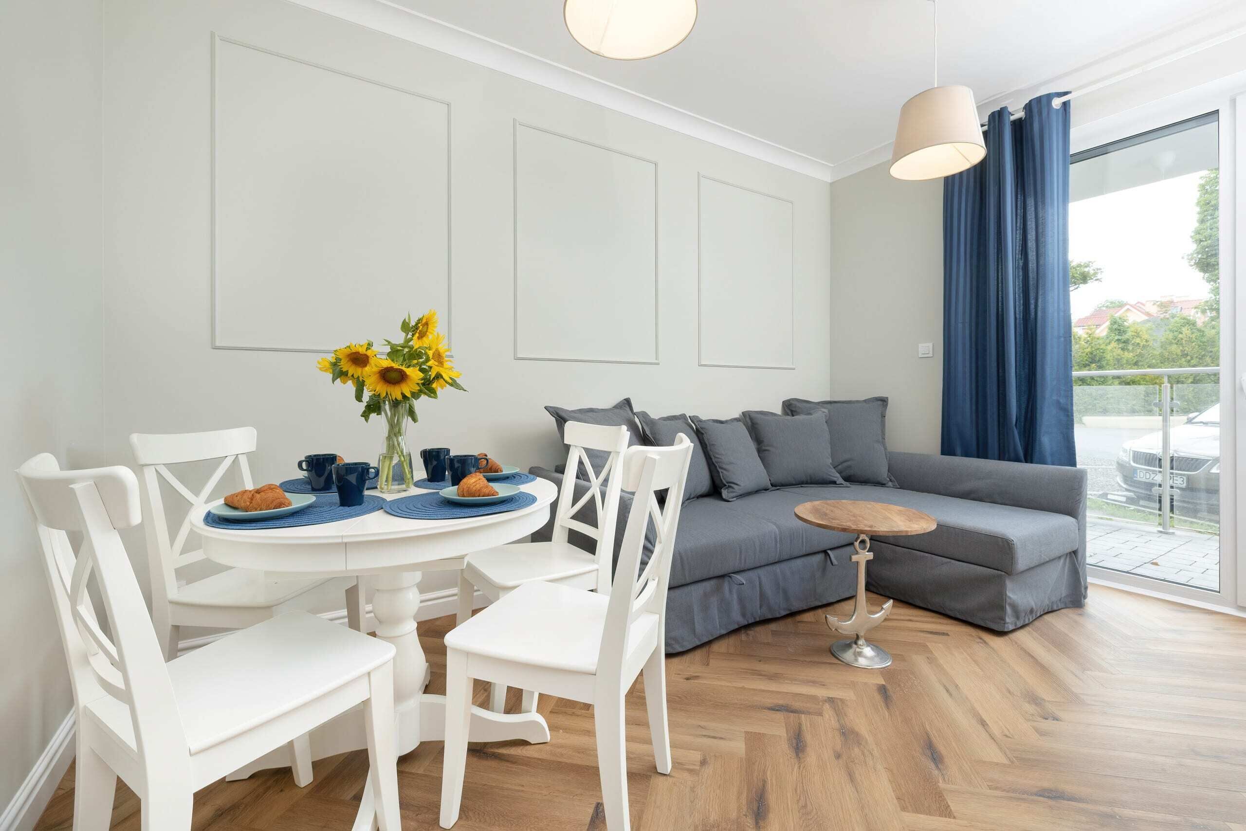 A bright dining and living area with a stylish gray sofa, blue accents, and a white dining set.

