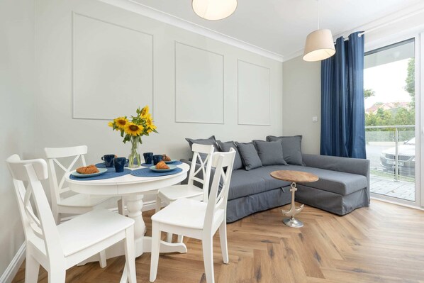 A modern living room with a round dining table, white chairs, a grey sofa, and a large window with blue curtains.