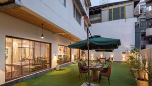 A courtyard with a dining area, artificial grass, potted plants, and a building with large windows and a balcony. - The Classio By DLS Hotels (Narendranagar)
