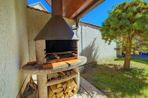 Ein an ein Gebäude angebauter, outdoor Holzofen, darunter ein Holzstapel und in der Nähe ein Baum.