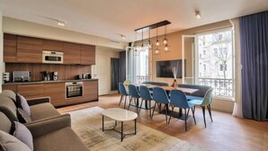 A modern living room with a sofa, a coffee table, and a dining area with chairs and a table. There is a kitchen with a microwave and a stove. - JARDIN DU LUXEMBOURG -RUE DE ASSAS -3 BEDROOM WITH AIR CON (paris)