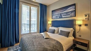 A hotel room with a large bed, a cityscape painting, a balcony with blue curtains, and a nightstand with a lamp. - JARDIN DU LUXEMBOURG -RUE DE ASSAS -3 BEDROOM WITH AIR CON (paris)