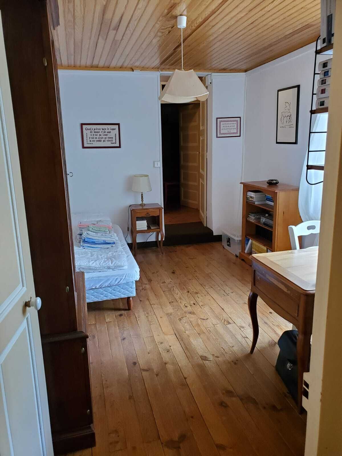 A small, cozy room with wooden floors, a bed, a desk, and a chair. There is a door leading to another room and a small table with a lamp.