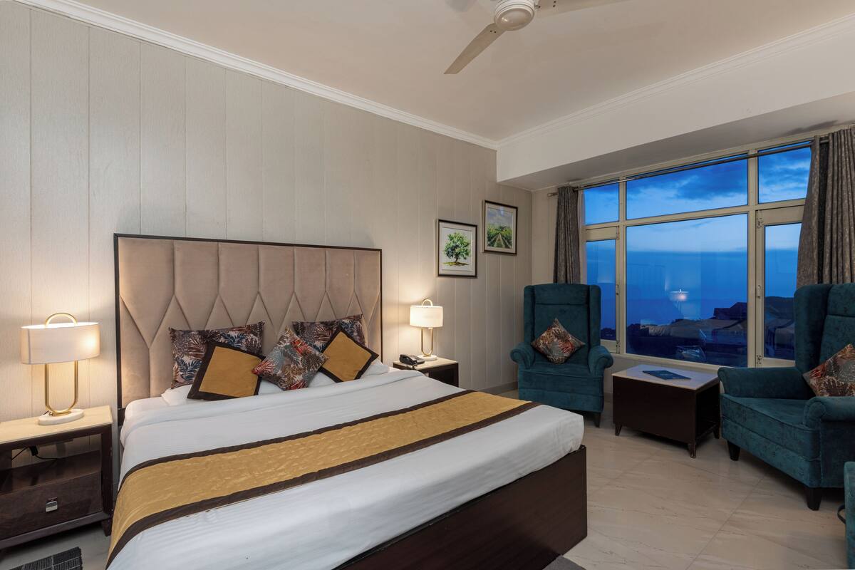 Habitación de hotel con una cama grande, dos sillas, una mesita pequeña, una mesita de noche y un ventanal con vista al océano.