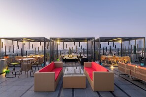 A rooftop terrace with wicker furniture, a central seating area, and a view of the cityscape. - Grand Luxotica By DLS Hotels (Dehradun)