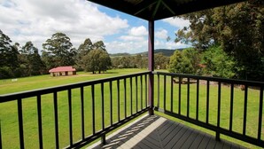 Un espacio al aire libre techado con una terraza de madera y barandilla de metal, con vista a un campo cubierto de césped y árboles.