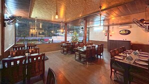 A restaurant with wooden tables and chairs, large windows, and hanging light fixtures. - MJ River Resort By DLS Hotels (Yamkeshwar)