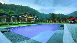 A pool area with a large rectangular pool, surrounded by buildings with a modern design, set against a backdrop of lush green hills. - MJ River Resort By DLS Hotels (Yamkeshwar)