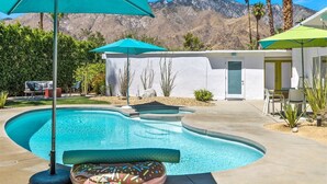 Una piscina con montañas al fondo, un edificio blanco con una puerta y un patio con sillas y sombrillas.