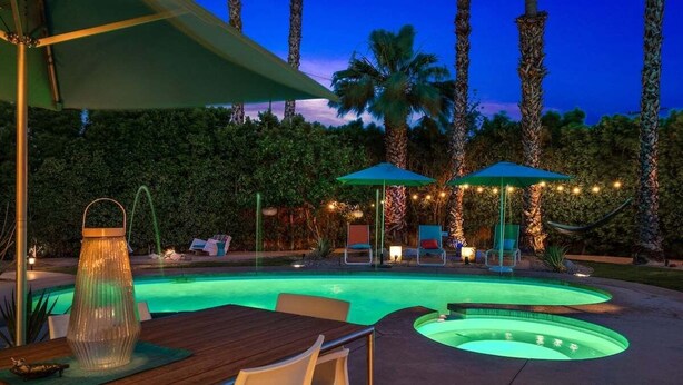 Una piscina de borde curvo, rodeada de palmeras y un patio con mobiliario exterior y luces colgantes.