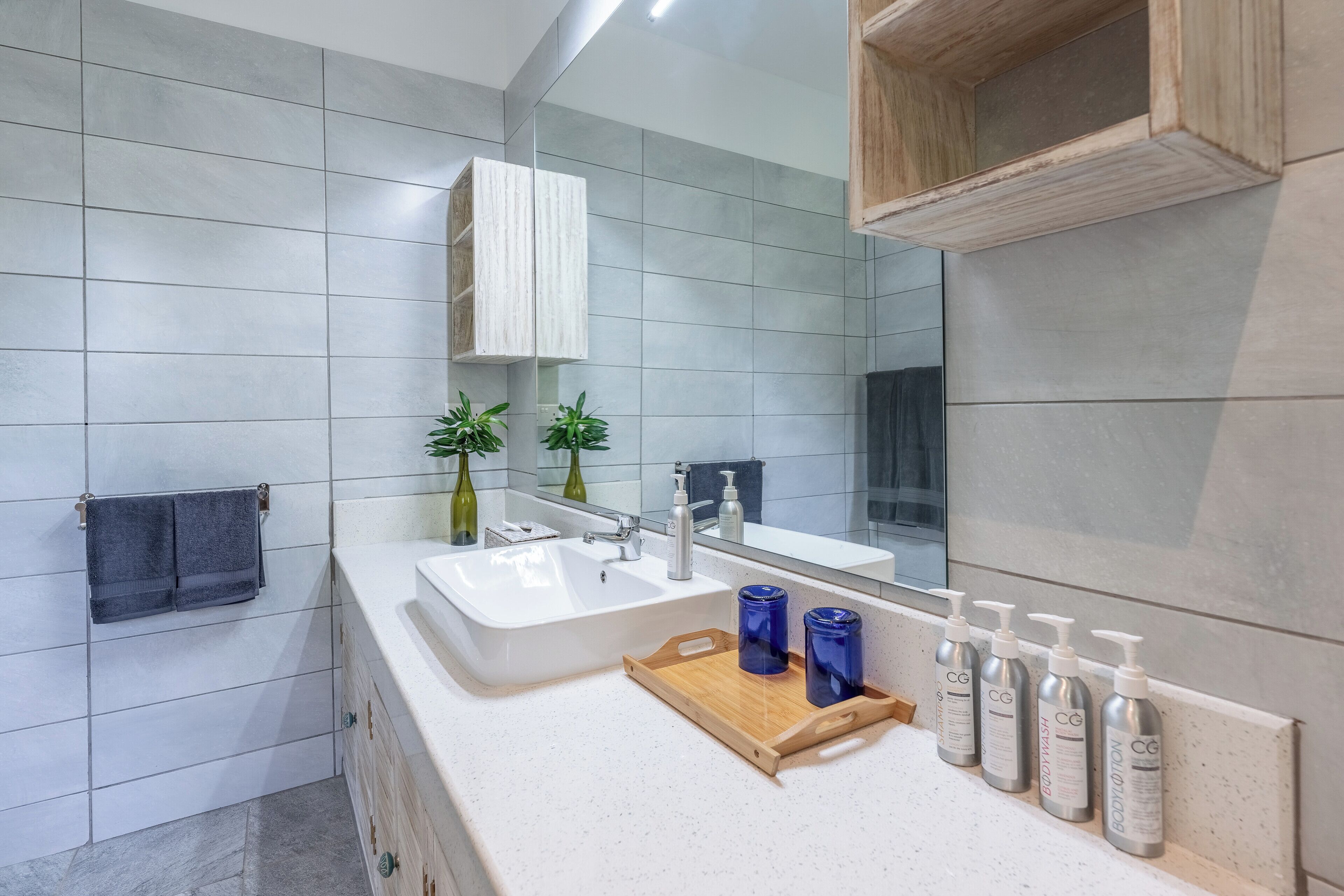 A modern bathroom with a large mirror, white tiles, a wooden vanity, and a tray with toiletries.