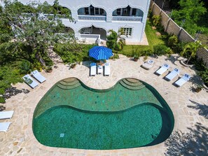 An aerial view of a swimming pool with lounge chairs and umbrellas, surrounded by a stone deck and lush greenery. - Hibiscus (Diani Beach)