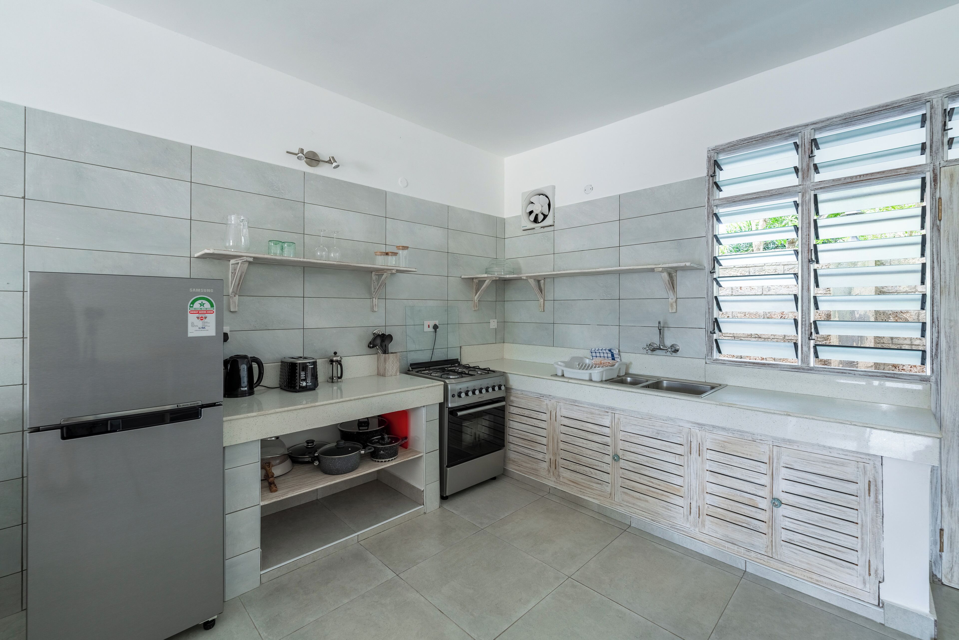 A modern kitchen with a stainless steel refrigerator, gas stove, oven, and white tiled backsplash. There are open shelves with kitchen items and a window with blinds.