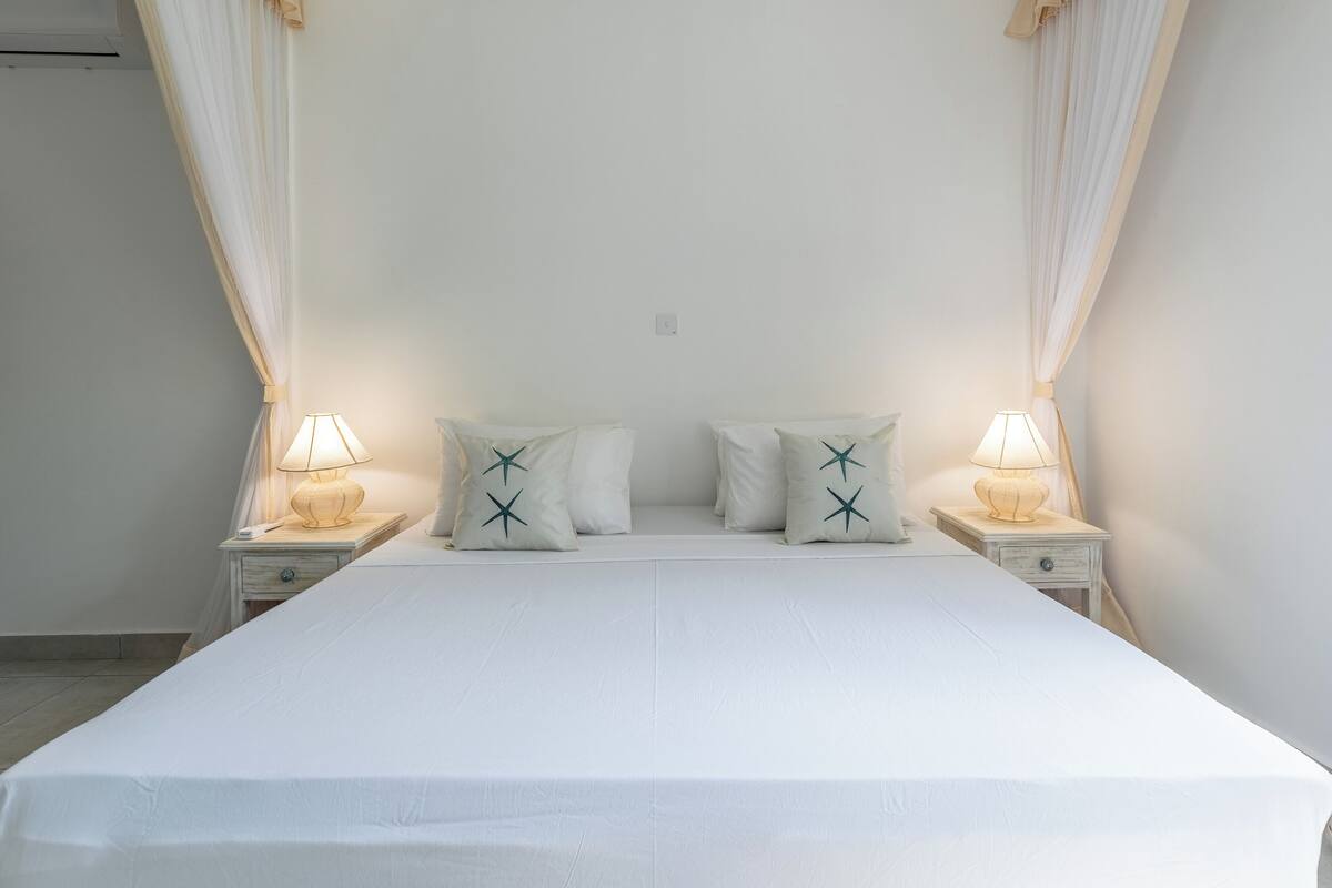 A neatly made bed with white bedding and pillows, two bedside lamps, and a canopy.