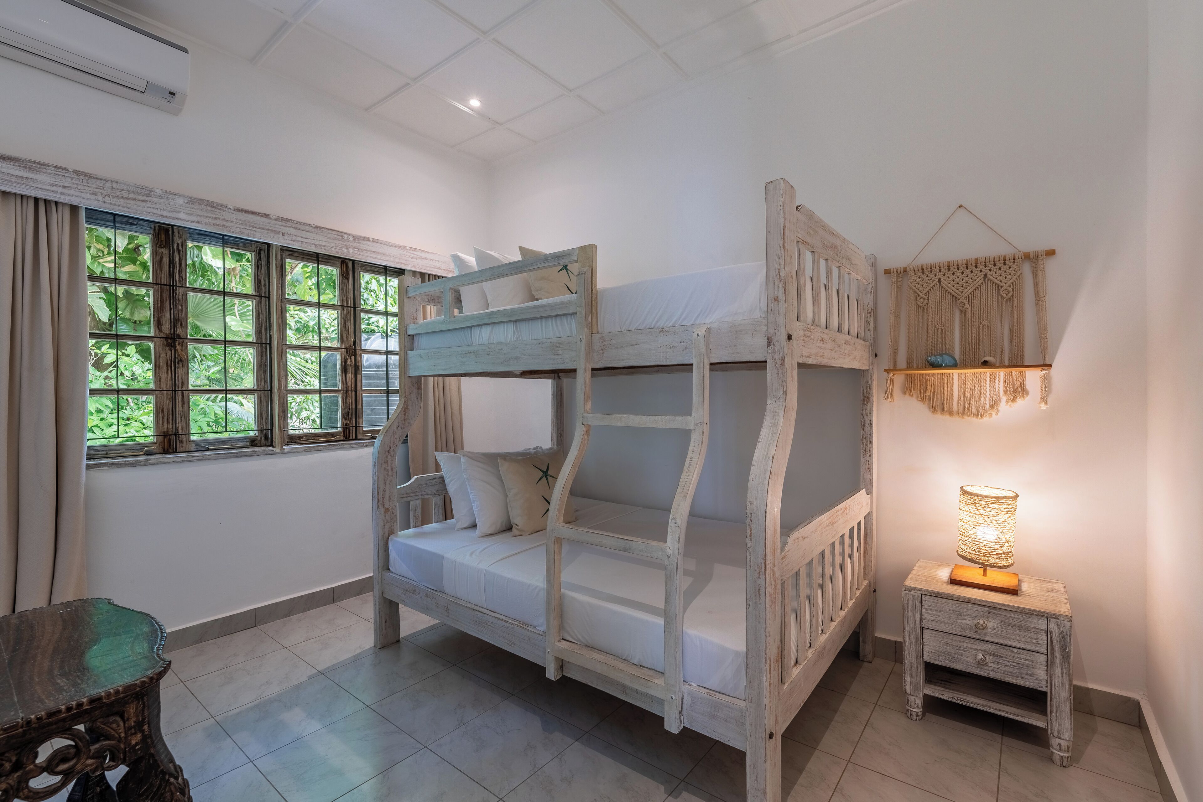 Una cama litera con una mesita de noche y una decoración de macramé colgante en una habitación con un ventanal grande.