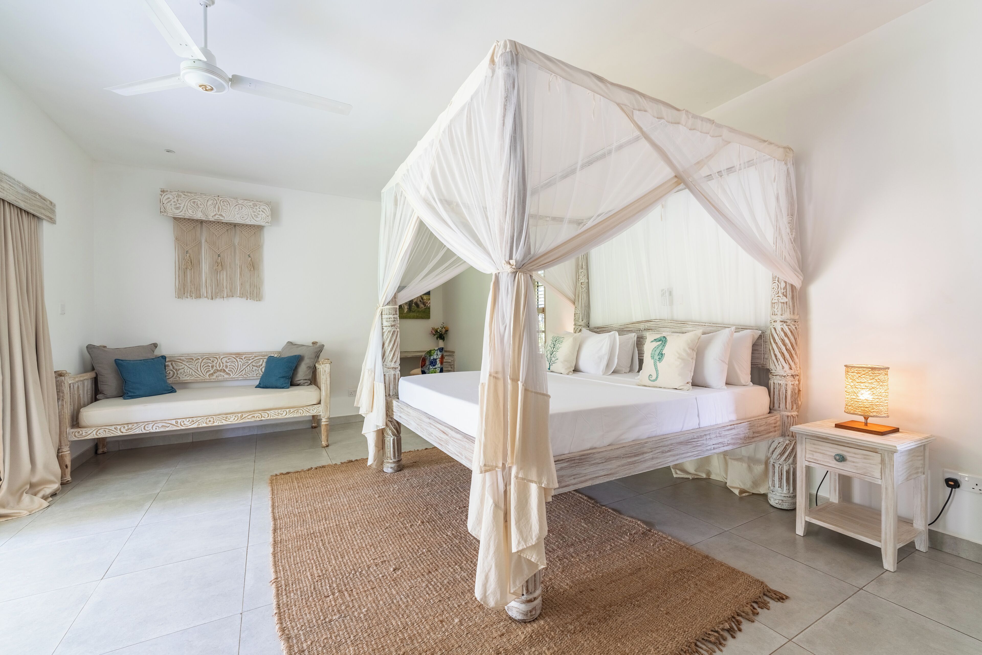 A bedroom with a canopy bed, a sofa, a bedside table with a lamp, and a ceiling fan.