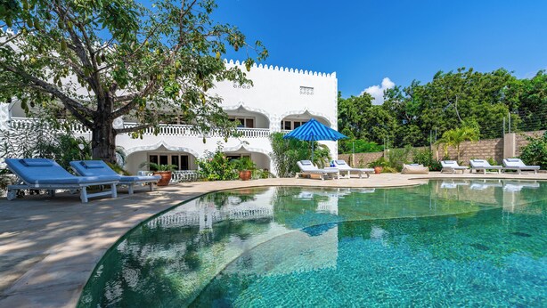 A clear swimming pool with lounge chairs and umbrellas, surrounded by a white building with balconies. - Hibiscus (Diani Beach)