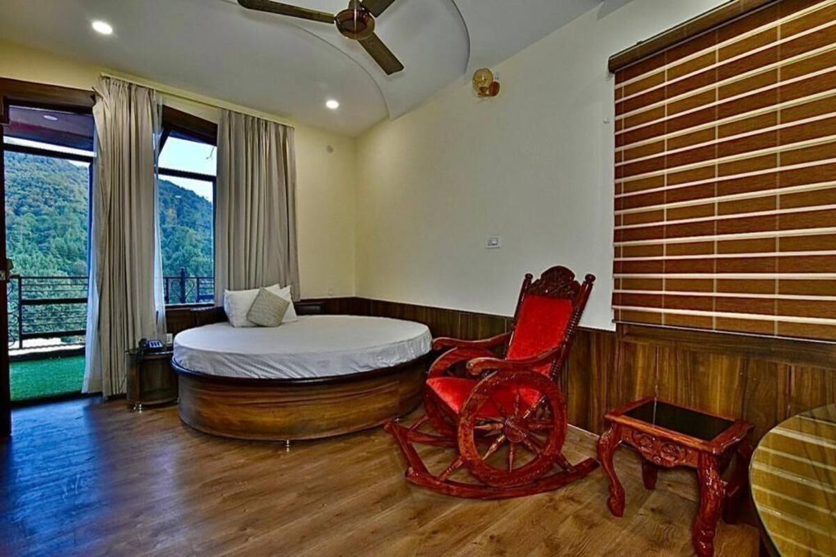 Une chambre à coucher avec un lit, un fauteuil à bascule rouge, une petite table, un ventilateur de plafond et une fenêtre avec des rideaux.